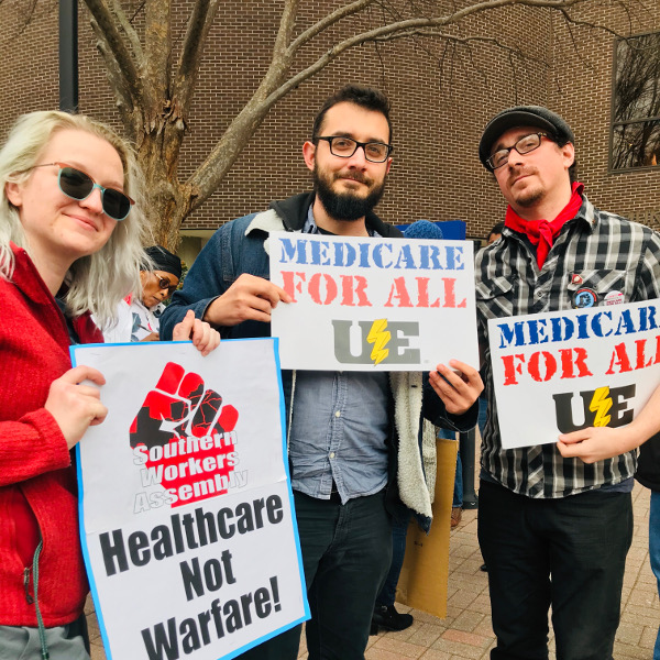 Young woman with Southern Workers Assembly Healthcare Not Warfare sign and two young men with Medicare for All UE signs