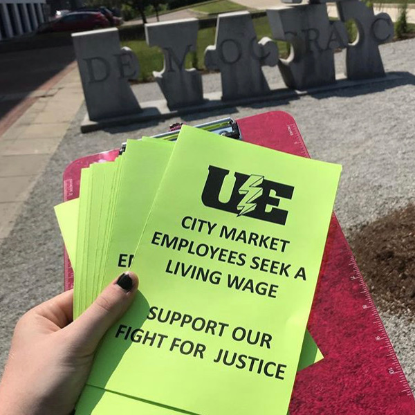 Union leaflets with a UE logo and the text "City Market employees seek a living wage. Support our fight for justice." Behind the leaflets is a sculpture of puzzle pieces that spell out the word "democracy."