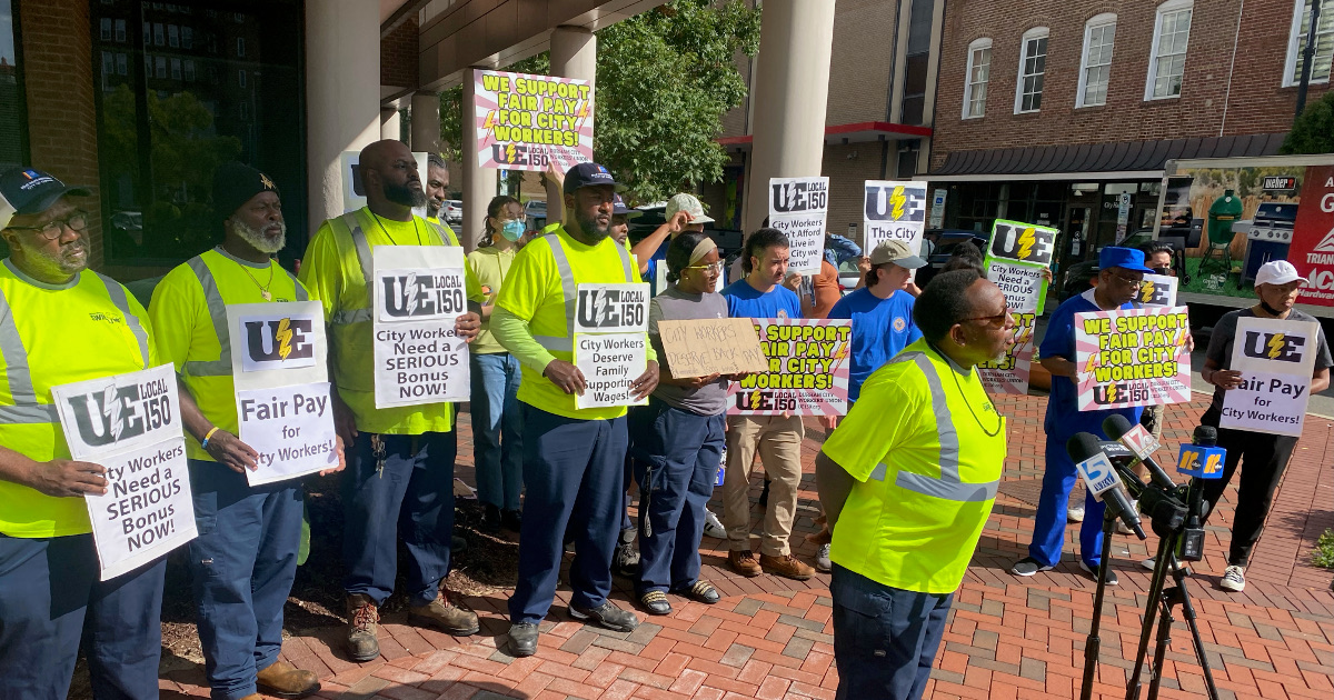 City workers with UE Local 150 signs demanding fair pay standing behind a worker at a microphone