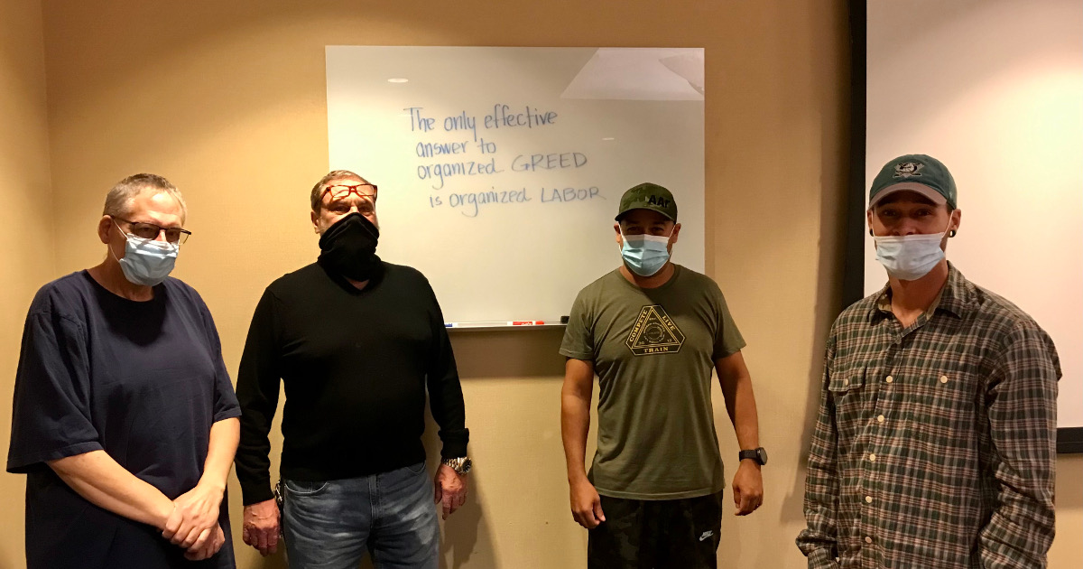 Four men wearing masks in front of a white board on which is written The only effective answer to organized GREED is organized labor.
