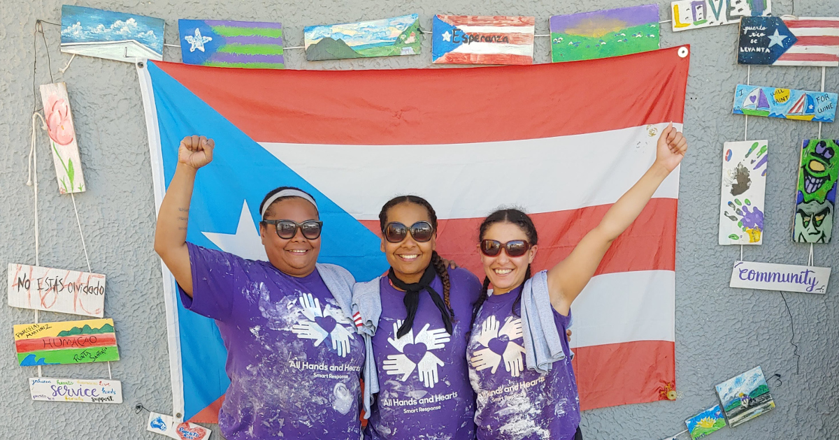 Three women in front of a Puerto Rican flag with raised fists and wearing purple t-shirts with a logo of four hands and the text "All Hands and Hearts Smart Response"