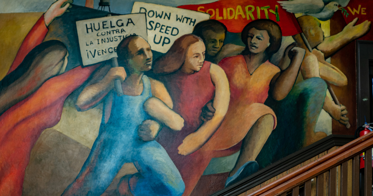 Image of a mural of a multiracial group of women and men with signs reading Solidarity, Huelga contra la injusticia, and Down with speed up