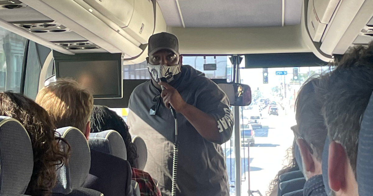 A Black man in a mask speaking into a microphone on a bus