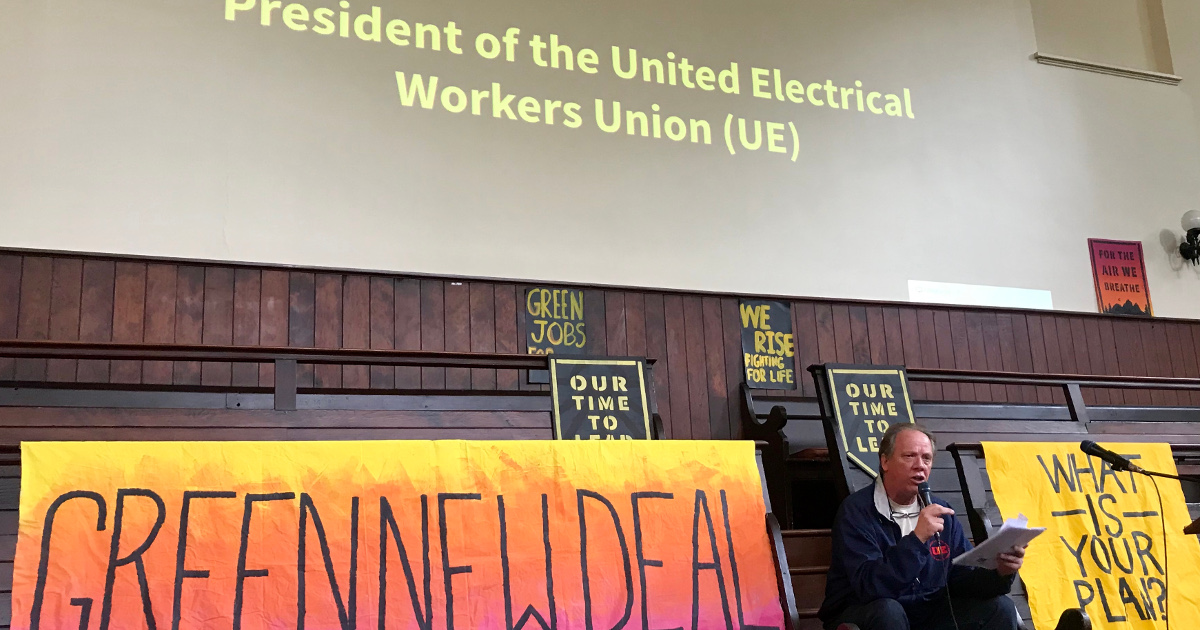 Speaker between banners reading "Green New Deal" and "What is your plan?" The words "President of the United Electrical Workers Union (UE)" are projected above the speaker.