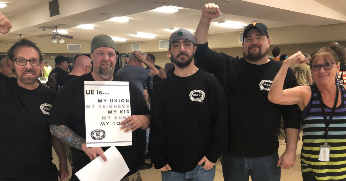 Five UE Local 610 members, four men and one woman, raising fists and holding a sign that reads "UE is ... my union, my neighbor, my kid, my aunt, my town, me."