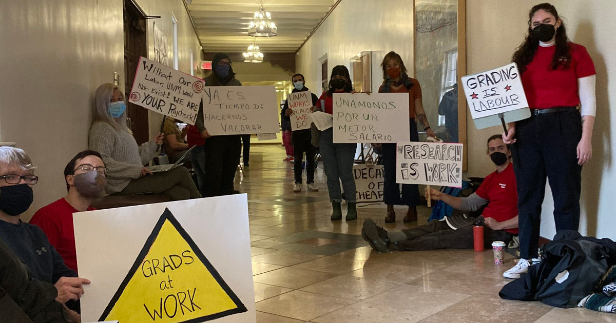UNM graduate workers in an academic building hallways with signs reading Grads at Work, Research is Work, Grading is Labor
