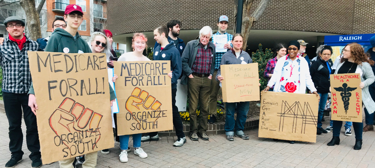Group of people with handmade cardboard signs reading "Medicare for All! Organize the South" and "Healthcare Is a Human Right"