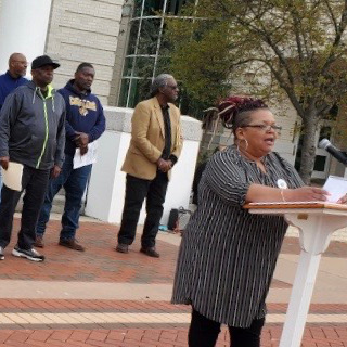 Black woman speaking into a microphone with four Black men behind her