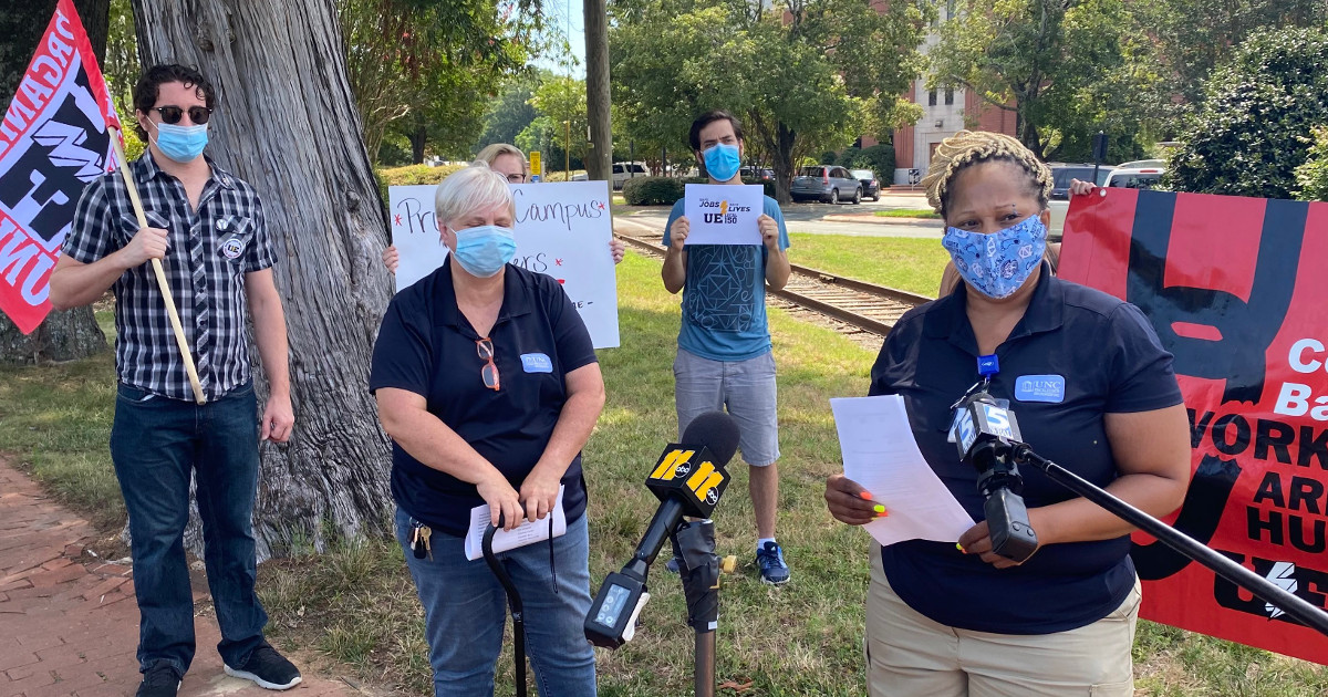 Woman worker in mask speaking into a microphone with other workers in masks with UE signs and flags