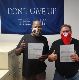 Two men wearing masks with thumbs up, standing in front of a banner reading Don't Give Up the Ship