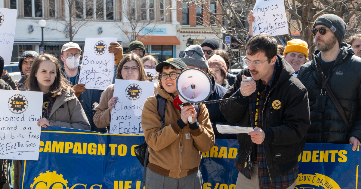 Cary Stough speaking in front of a group of grad workers with signs indicating different departments opposed to the fees.