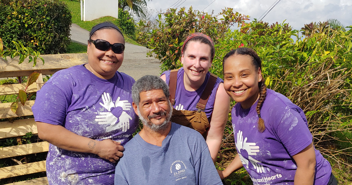 Three women wearing purple "All Hands and Hearts Smart Response" t-shirts behind an older, seated man