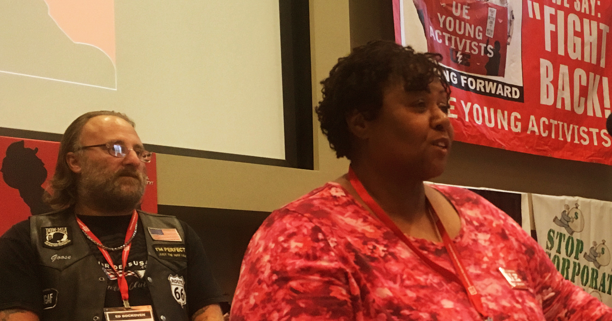 Photo of an African-American woman speaking and a white man standing behind her (to the left)