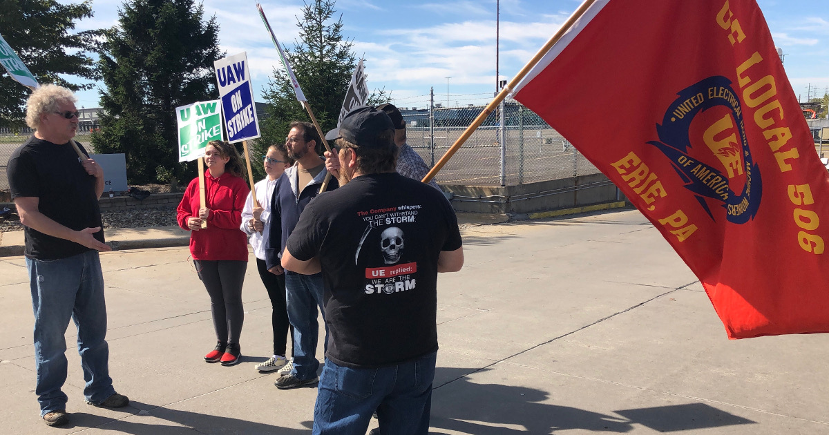 UE Local 506 members speaking with a small group of GM strikers. One of the UE 506 members holds a large red flag with a UE logo and the words "UE Local 506, Erie, PA"