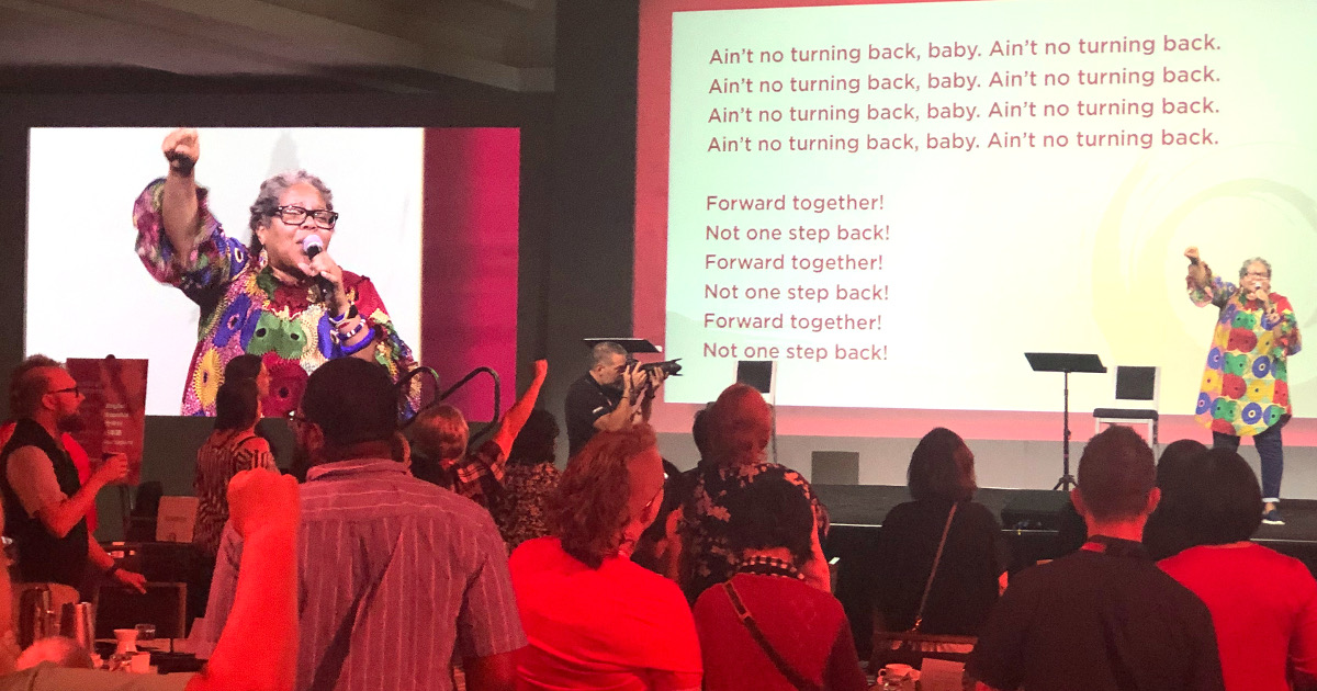 Nathanette Mayo singing in front of a group. Behind her on the screen are the words "Ain't no turning back, baby. Ain't no turning back" (repeated four times) and "Forward together! Not one step back!" (repeated three times)