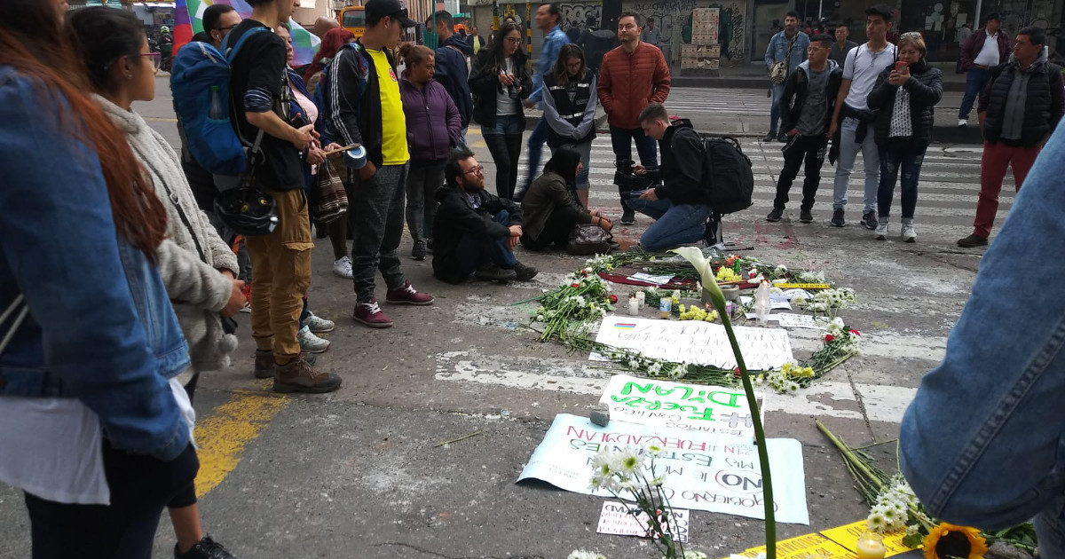 A group of people gathered around a memorial of signs and flowers laid out on a street