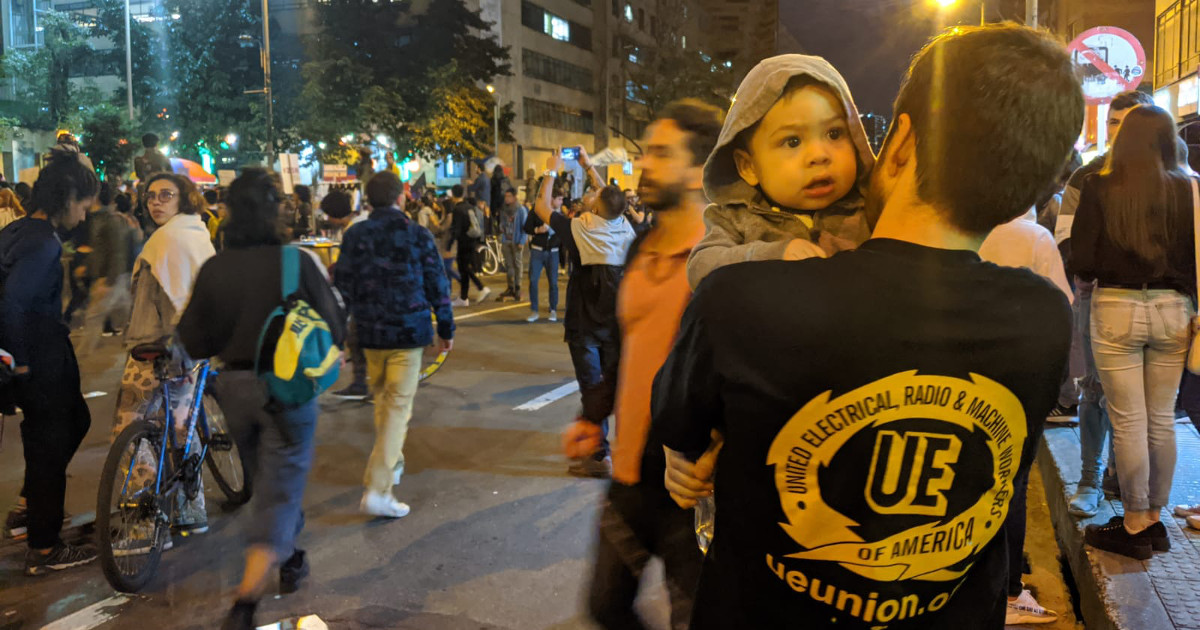 Crowd in the street at night. At right front of the photo, a young man holds a baby. His back is to the camera, displaying a t-shirt with a large UE logo on it.